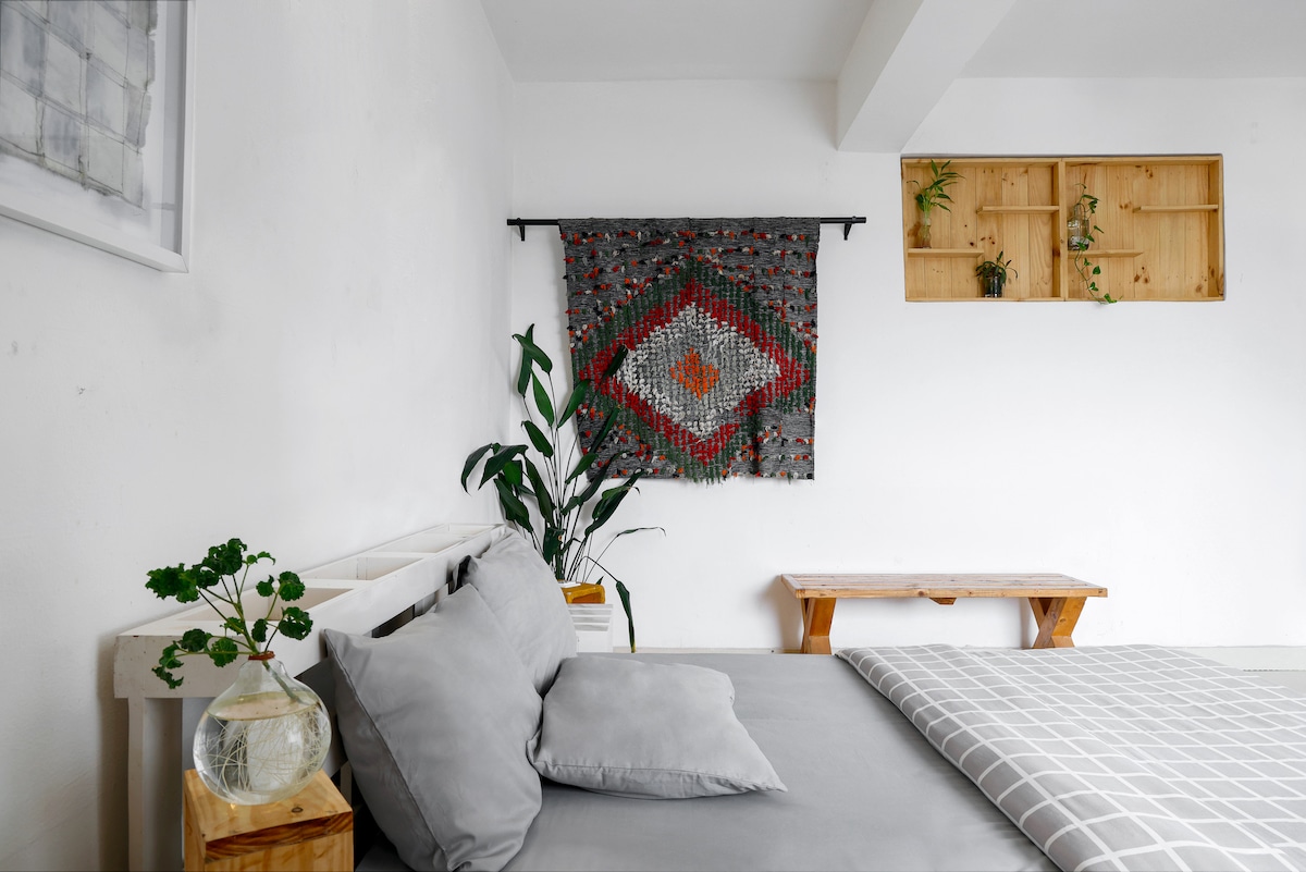 A serene room features a twin-size bed dressed in soft gray linens. A woven textile art piece hangs above the bed, adding color to the neutral walls. A small indoor plant is positioned beside the bed, and a light wooden table is visible across the space.