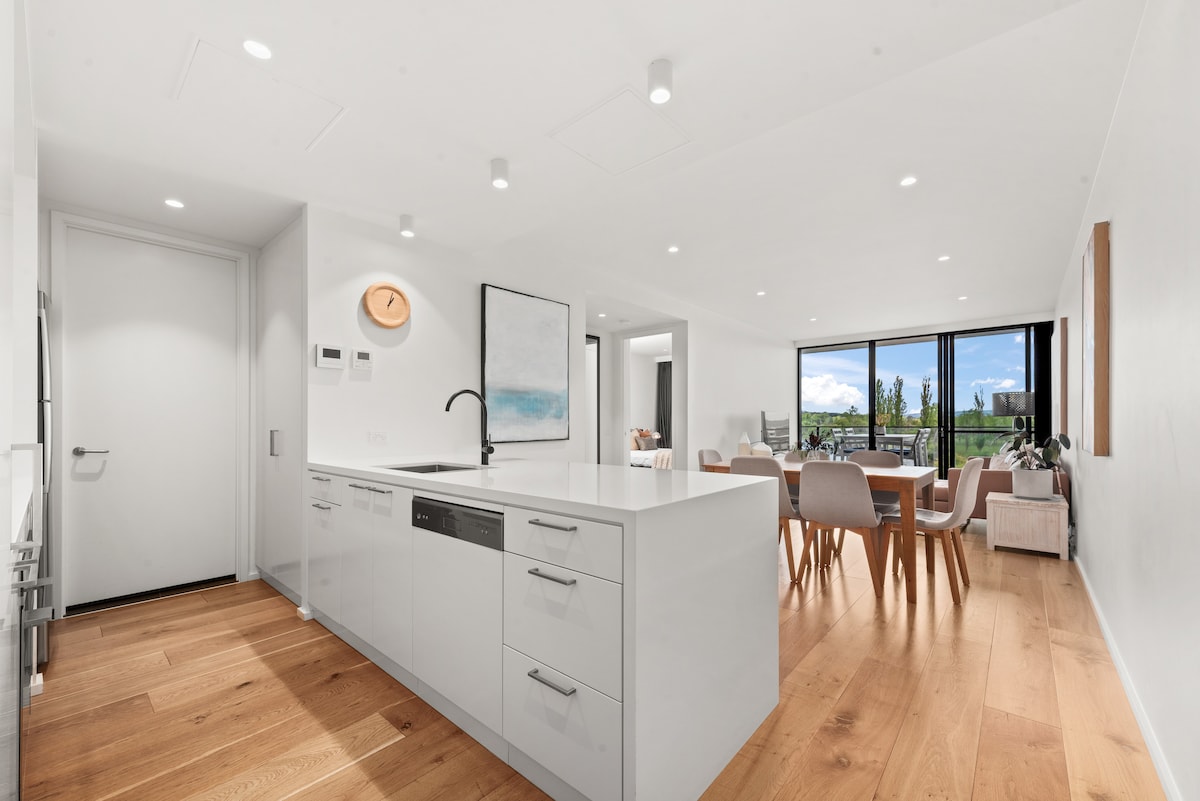 A modern kitchen is showcased with sleek white cabinetry and a polished countertop. Natural light floods the open dining area, featuring a wooden table surrounded by stylish chairs. An inviting living space with large windows offers views of outdoor greenery.
