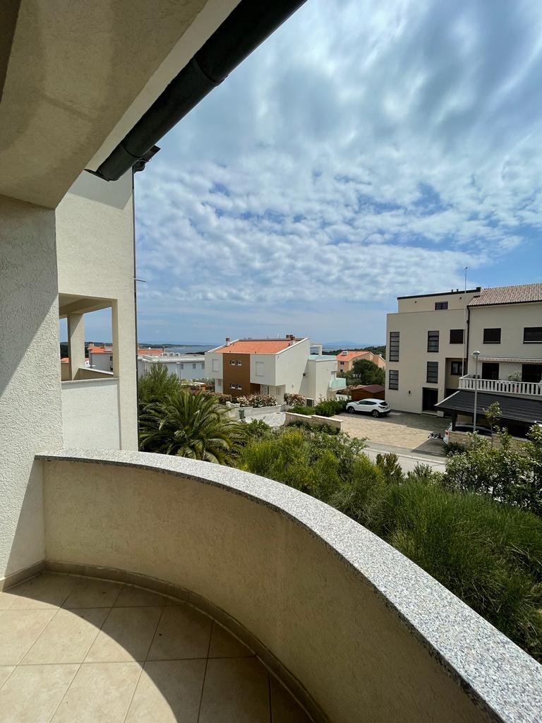 A view from the balcony presents a landscape of neighboring buildings and glimpses of the sea in the distance. The curved balcony edge frames the scene, while palm trees and greenery are visible in the foreground against a backdrop of a partly cloudy sky.