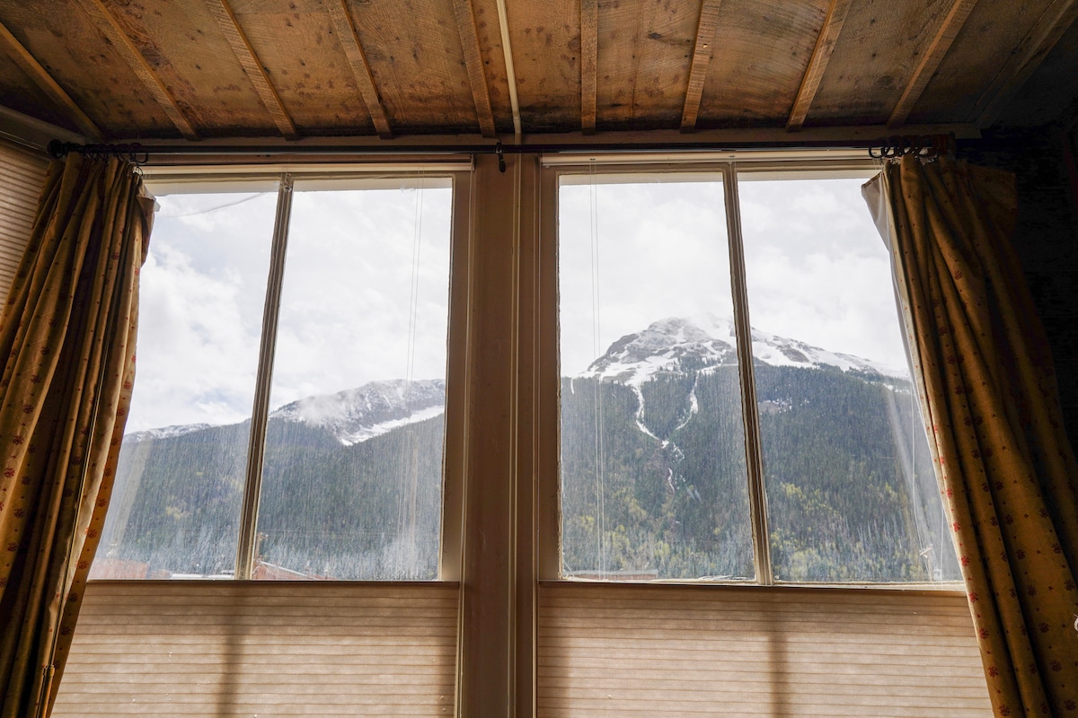 Two large windows frame a breathtaking view of snow-capped mountains and lush green forests. Natural light fills the space through the partially opened window coverings, creating a soothing atmosphere within the room.