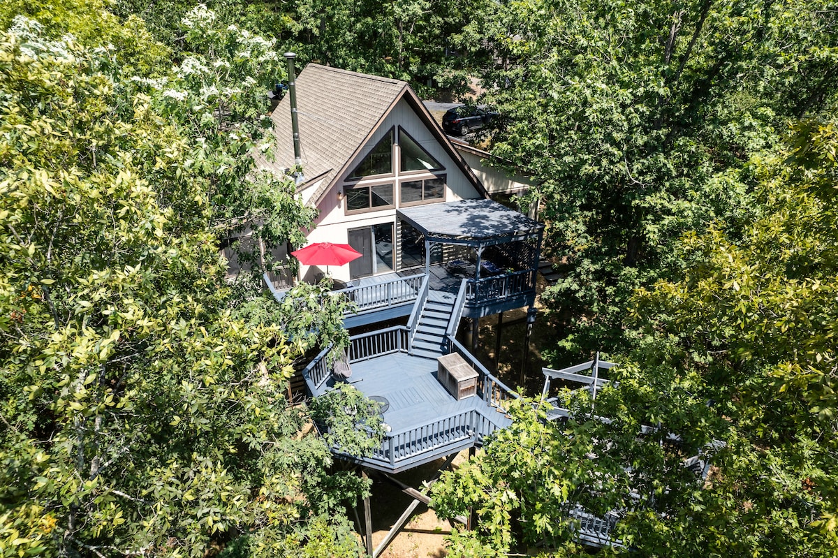 A two-story house nestled among trees features large outdoor decks. A red umbrella is positioned on the lower deck, offering a pop of color. Expansive windows allow natural light to flood the interior, creating a connection with the surrounding nature.