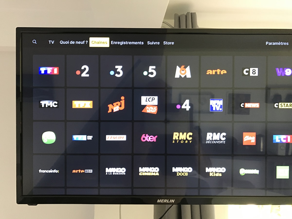 A television screen displays a grid of channels, showcasing various logos. The interface includes options for live TV, recordings, and a store. The background is a soft grey, with curtains visible beside the television.