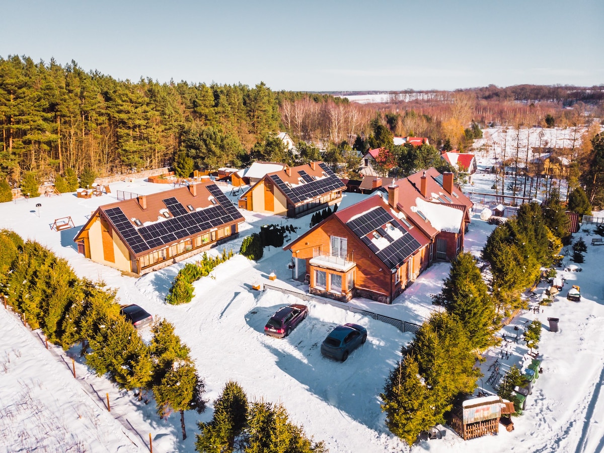 A view of the Malinowy Dworek complex reveals several buildings surrounded by a snowy landscape. Solar panels adorn the rooftops, and a driveway accommodates parked cars. Adjacent green trees and open space enhance the tranquil, winter setting.