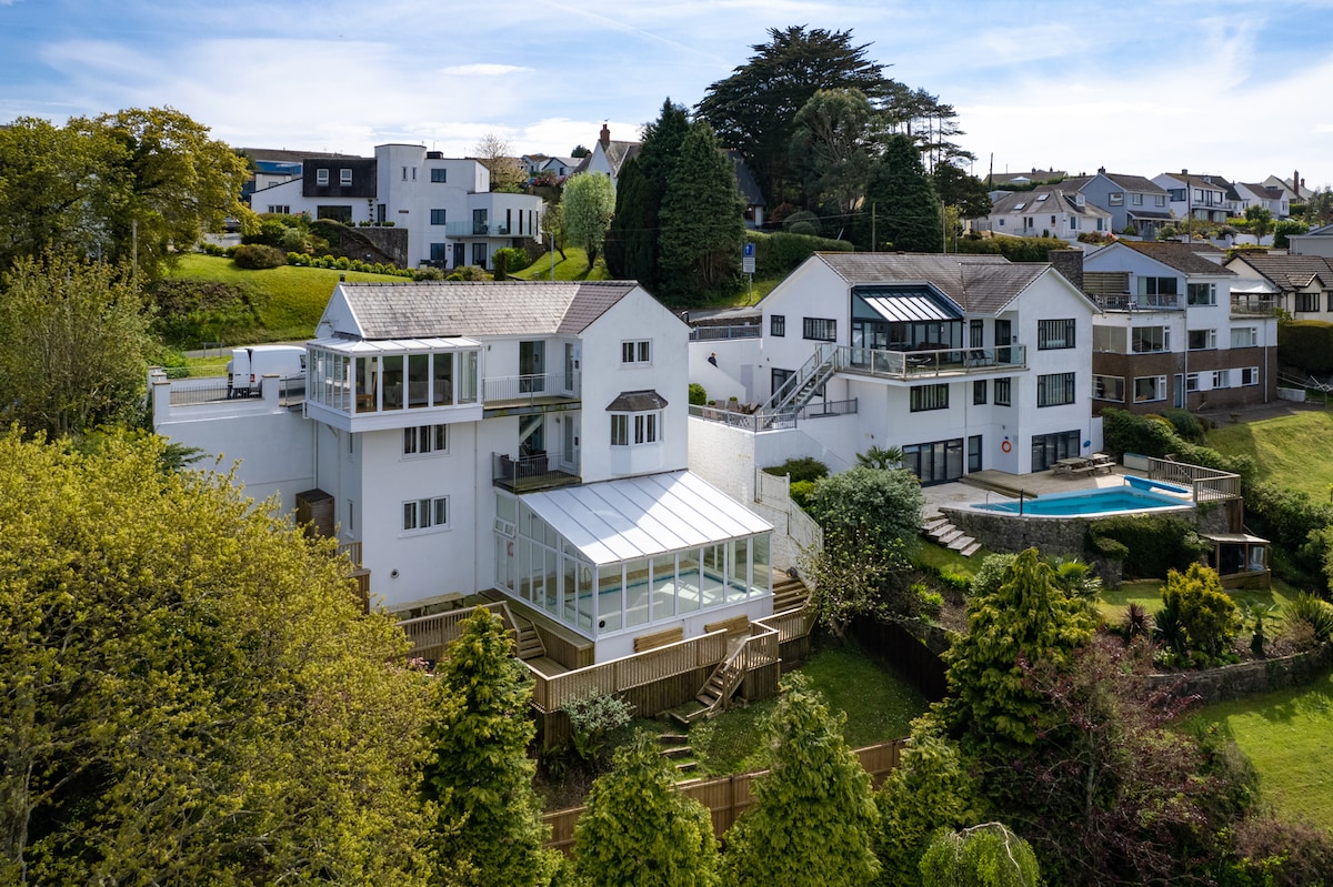 A multi-level house is shown, surrounded by lush greenery. Large windows and light-colored walls reflect a modern design. A private swimming pool is visible in the backyard, complemented by outdoor seating areas, enhancing the outdoor living space. Scenic views of the landscape can be seen in the background.