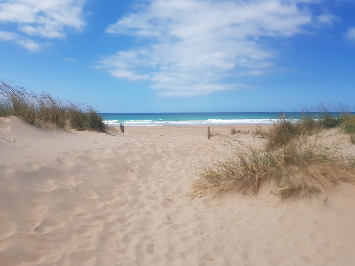 The sandy path leads through coastal vegetation to a serene beach with gently rolling waves. The horizon features a clear blue sky, while the ocean displays various shades of turquoise, creating a peaceful coastal scene.