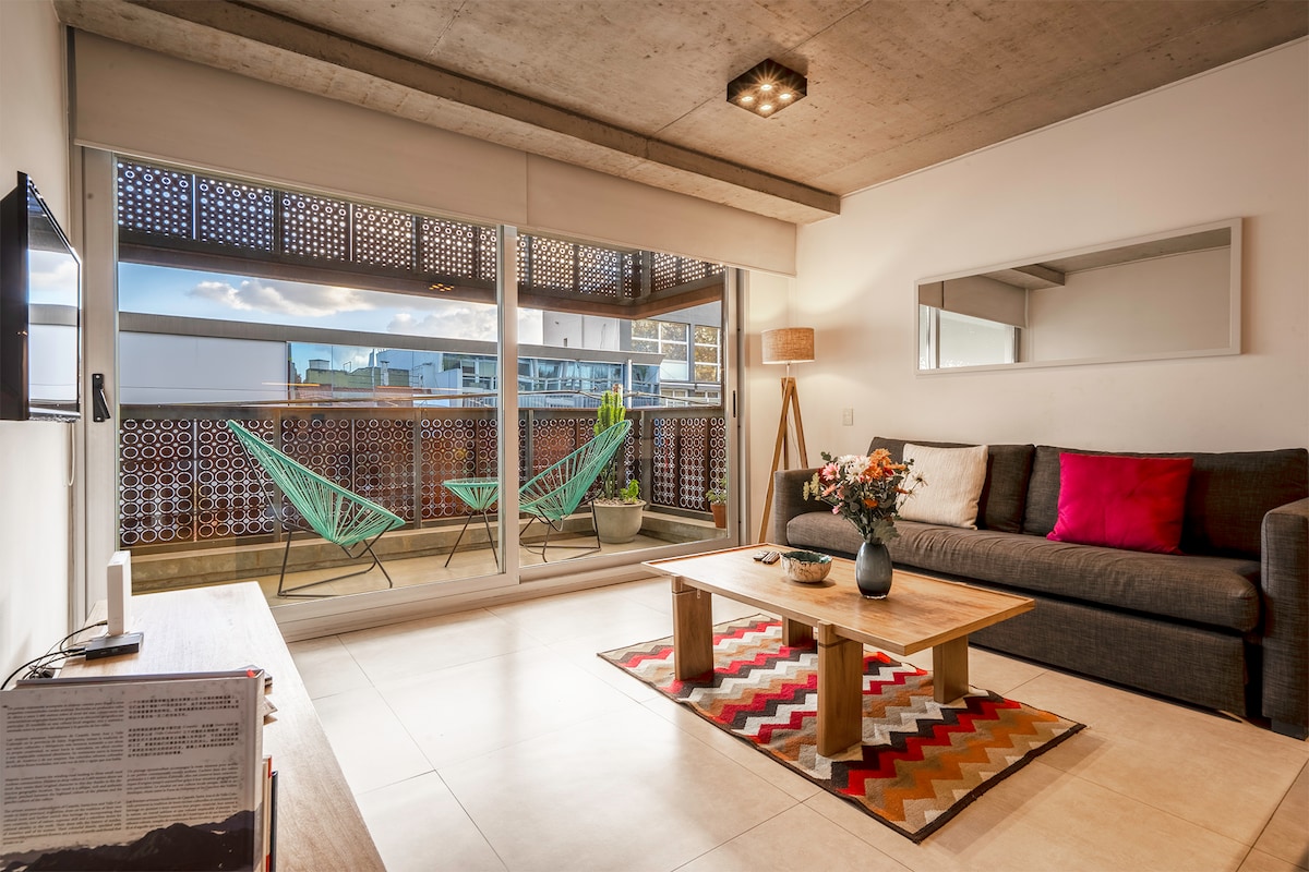 A modern living area features large glass doors that open to a balcony, allowing natural light to fill the space. A comfortable gray sofa is complemented by decorative pillows and a coffee table. A patterned rug adds color, while potted plants and lounge chairs enhance the outdoor feel.