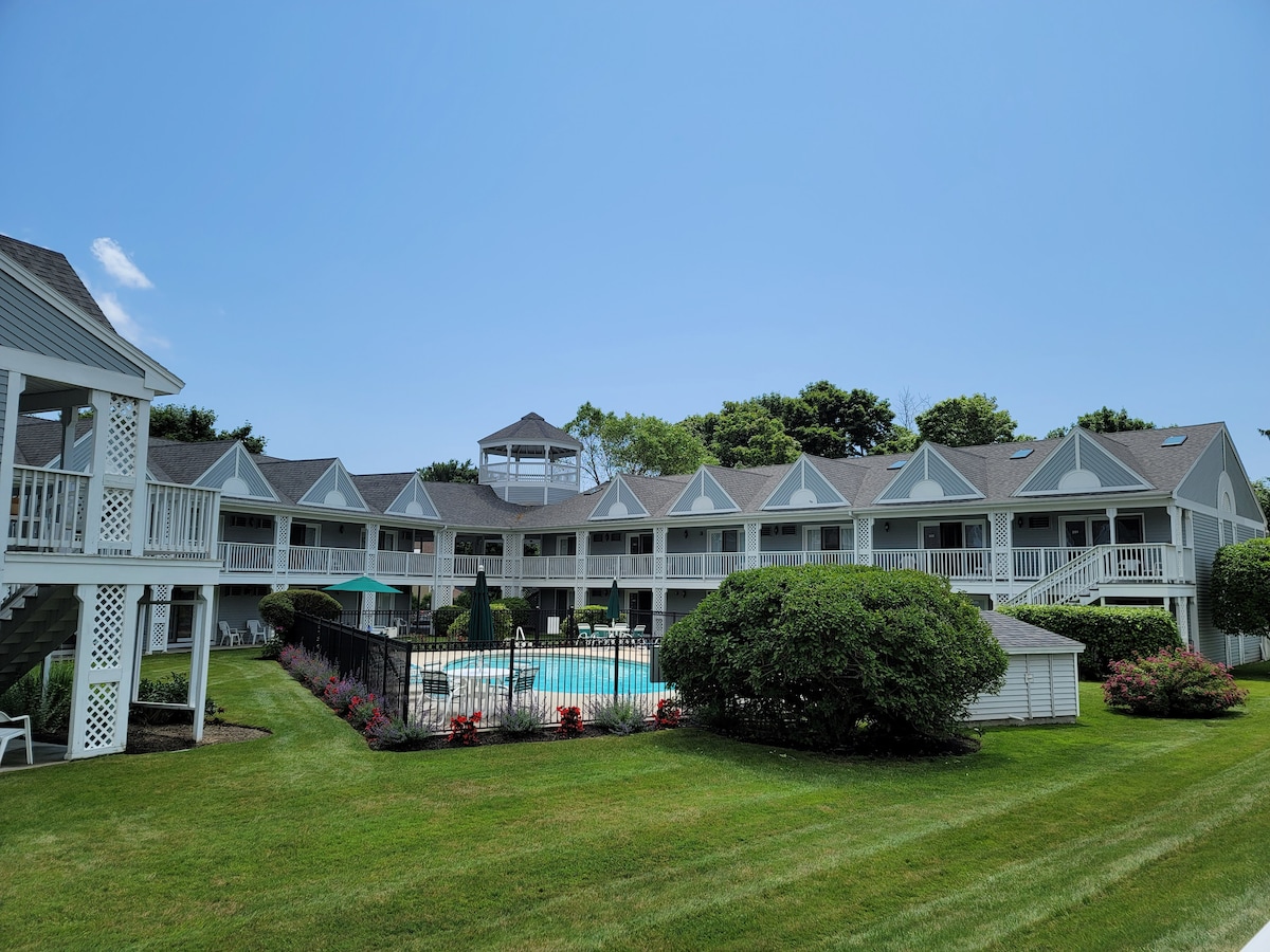 The building features a two-story design with a view of the well-maintained pool area surrounded by manicured lawns and colorful flower beds. Clear blue skies provide a bright backdrop for the structure, and shaded sitting areas can be seen near the pool.