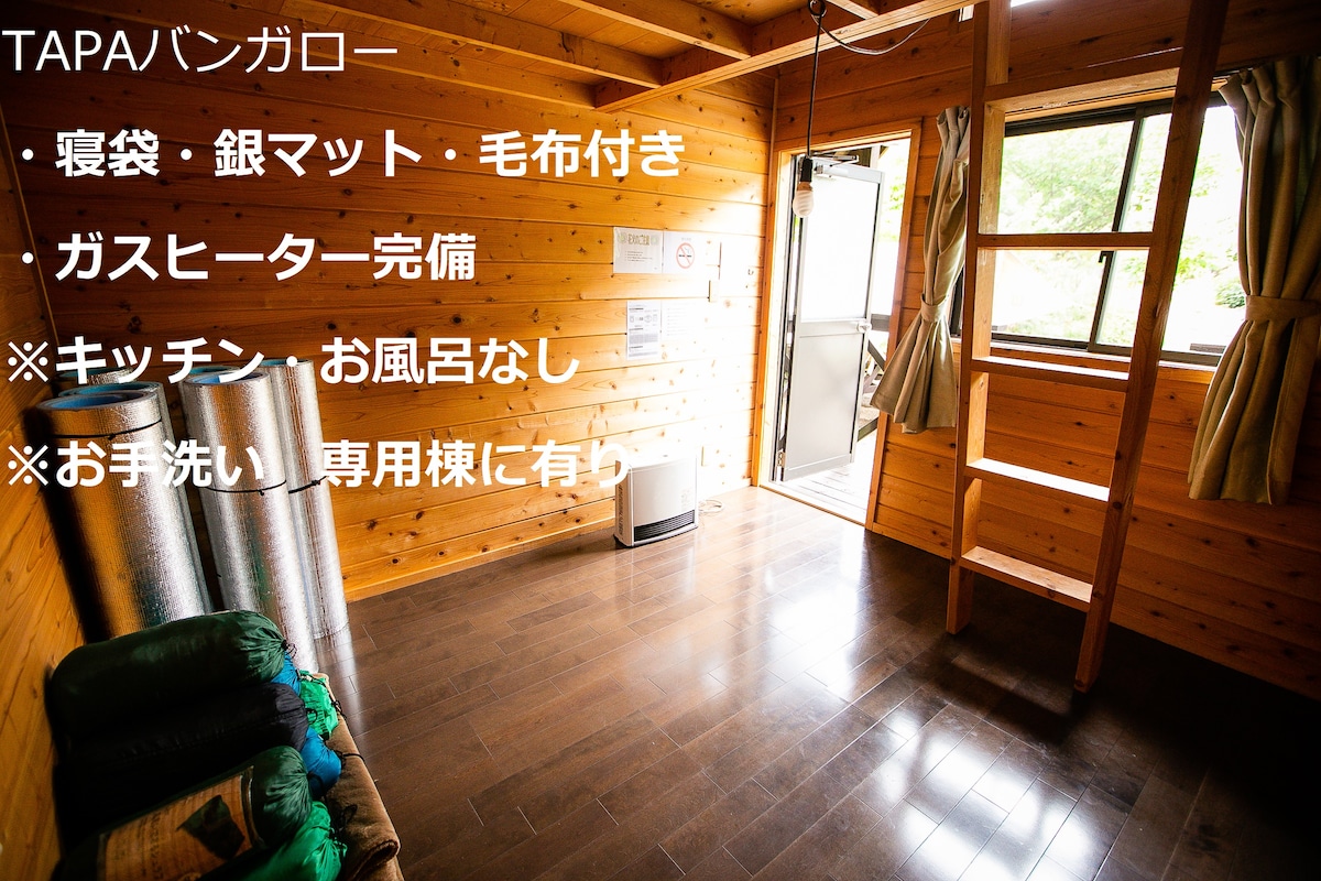 The interior of a wooden bungalow showcases natural wood walls and flooring. A gas heater is positioned unobtrusively, while sleeping bags and silver mats are provided. A window offers natural light, with views of the outdoors visible, creating an open and inviting space.