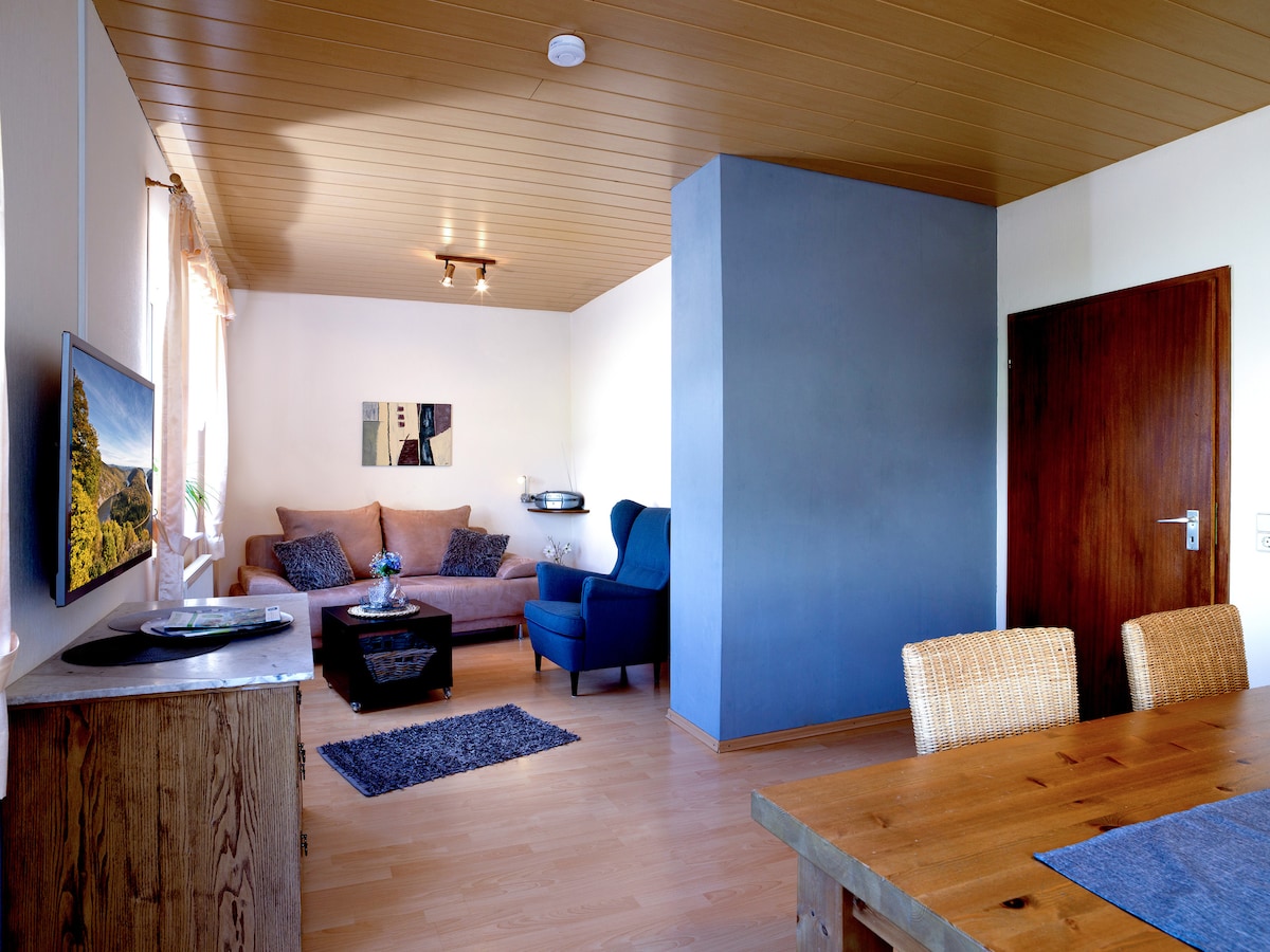 The living area features a cozy seating arrangement with a sectional sofa and an armchair, complemented by a wooden coffee table and a flat-screen TV. A dining table with woven chairs occupies the space, and natural light enters through a window, illuminating the room.