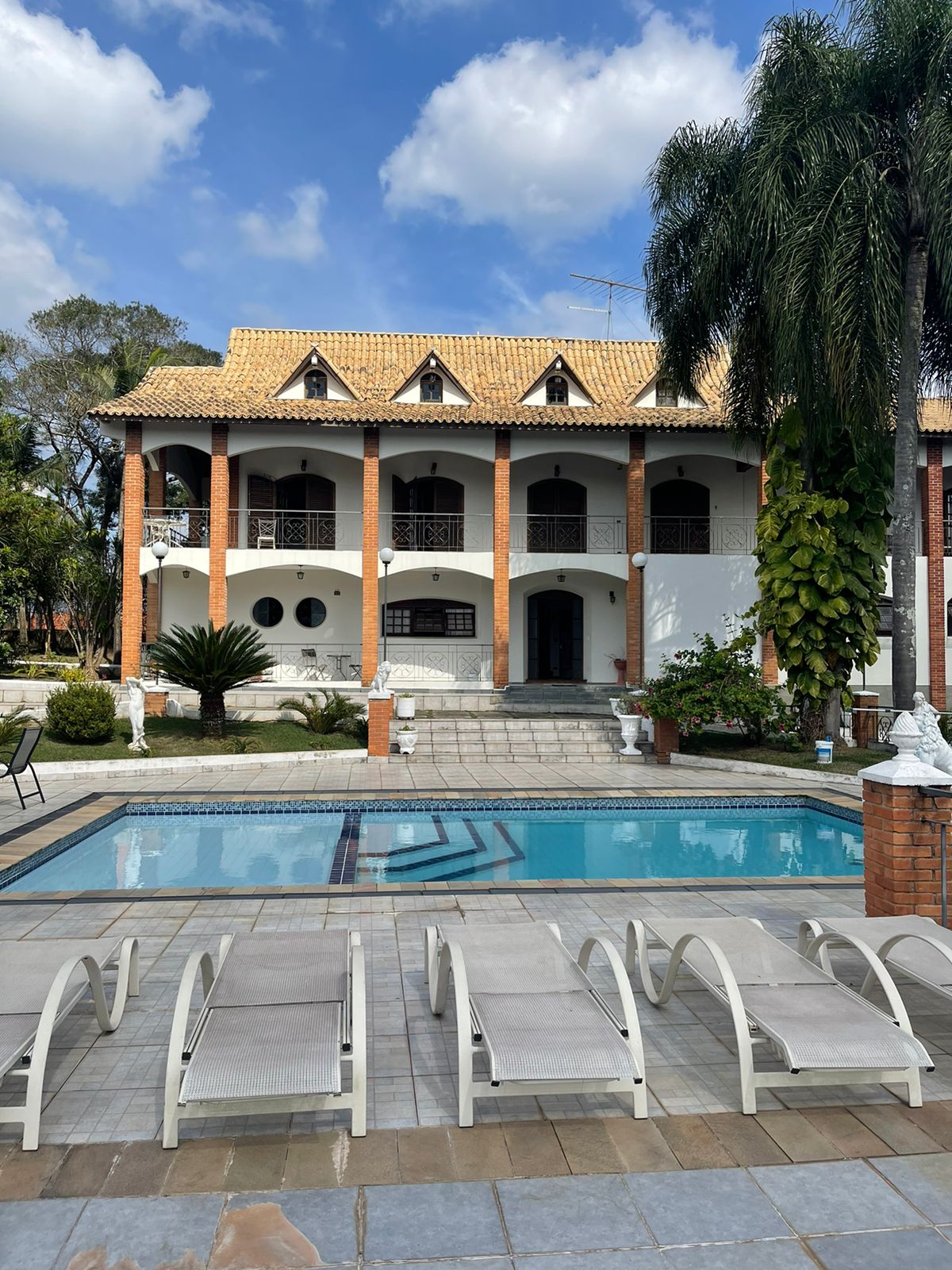 The exterior of a spacious three-story house is presented with a tiled roof and elegant arches. A clear swimming pool is framed by lounge chairs, surrounded by lush greenery and well-maintained gardens, offering a welcoming outdoor environment.
