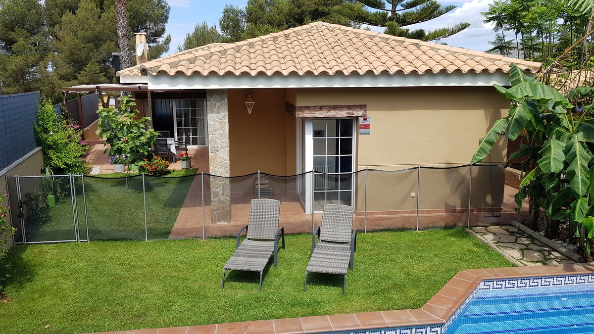 A single-story house is displayed, featuring a light-colored façade and a tiled roof. Lounge chairs are positioned on well-maintained grass near a swimming pool, framed by lush greenery. A safety fence encloses the pool area, ensuring a secure environment for families.