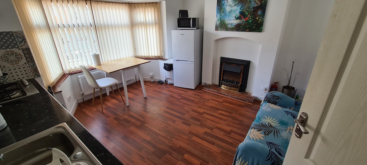 A bright kitchen/living area features a wooden dining table with a white chair, a refrigerator, and a microwave. The room is accented by a cozy sofa and a decorative fireplace. Natural light streams through the large bay windows covered by vertical blinds.