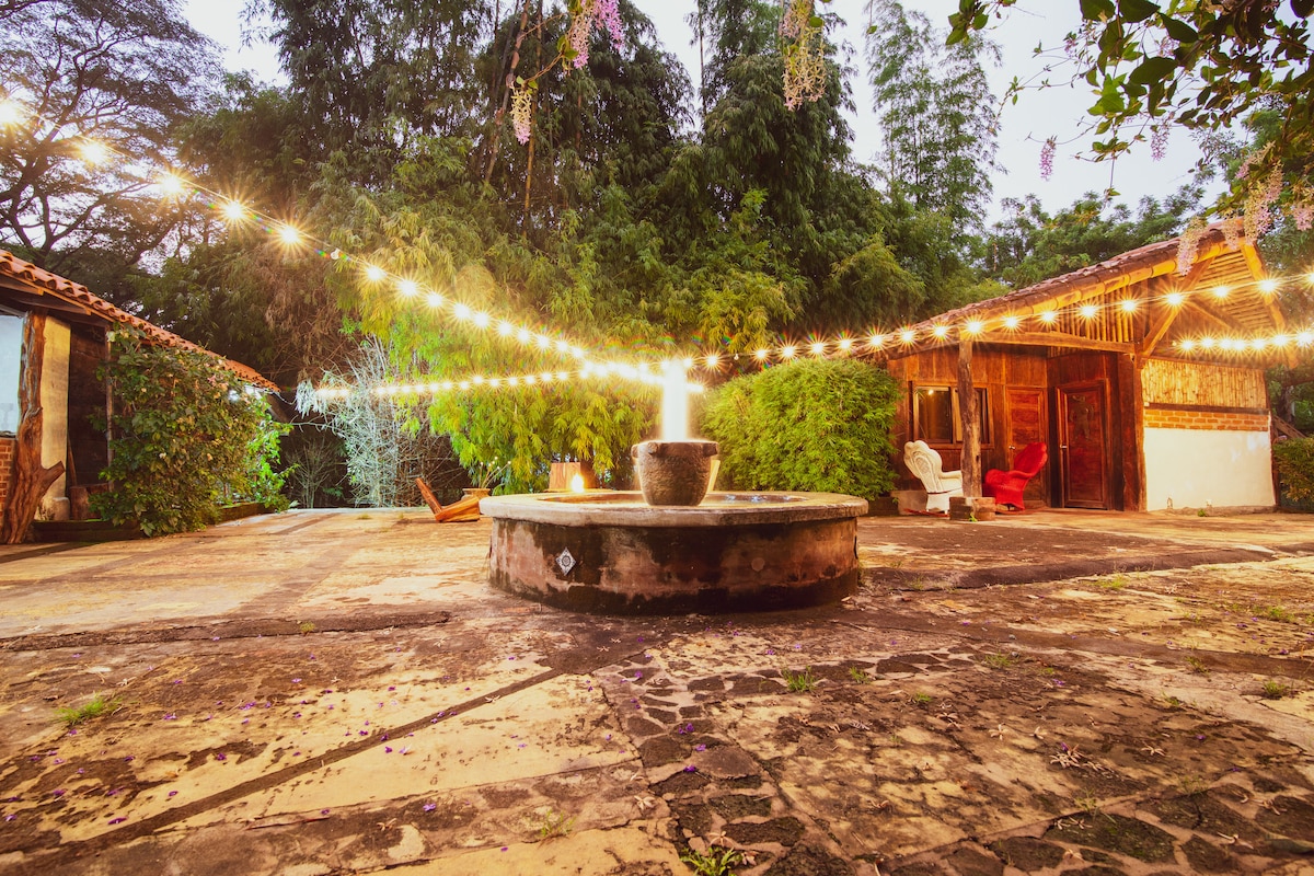 A peaceful outdoor area is illuminated by string lights, creating a soft glow over a circular stone fountain. The surrounding space features rustic buildings and lush greenery, providing a serene setting for relaxation and gatherings.