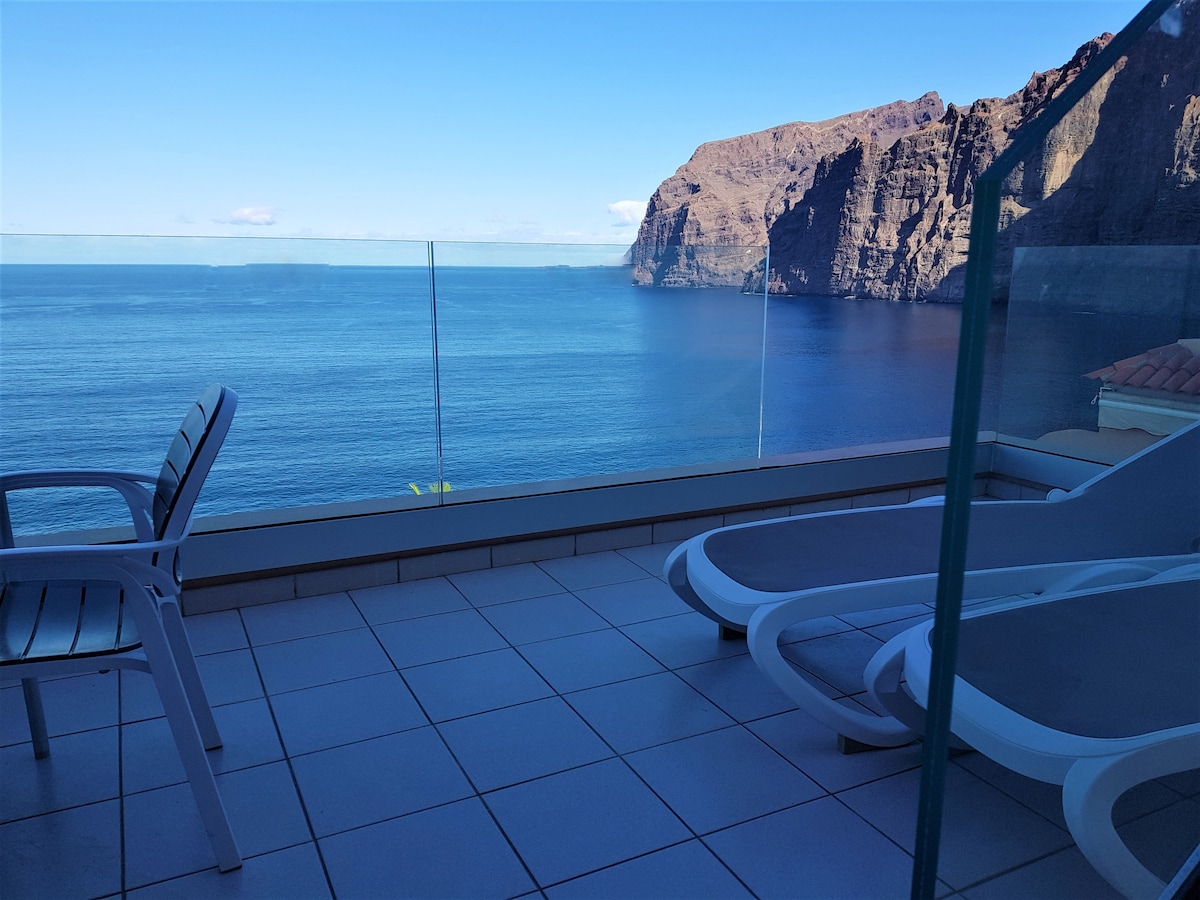 A terrace provides unobstructed views of the sea and the striking cliffs in the distance. White lounge chairs sit adjacent to a glass railing, creating a seamless connection to the surrounding landscape. The tranquil water reflects the clear blue sky above.
