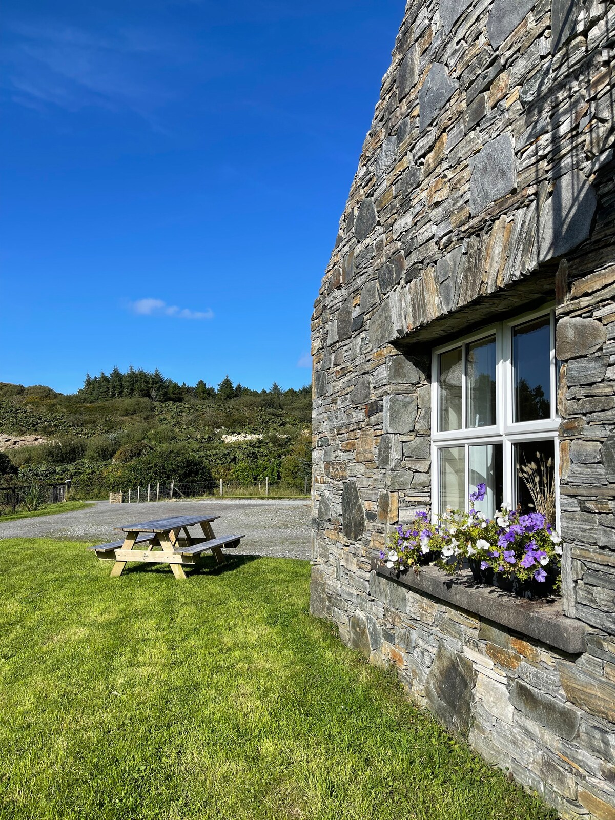 A rustic stone building is depicted, framed by a large window adorned with blooming flowers. A wooden picnic table rests on a grassy area in front, surrounded by trees and hills against a bright blue sky.