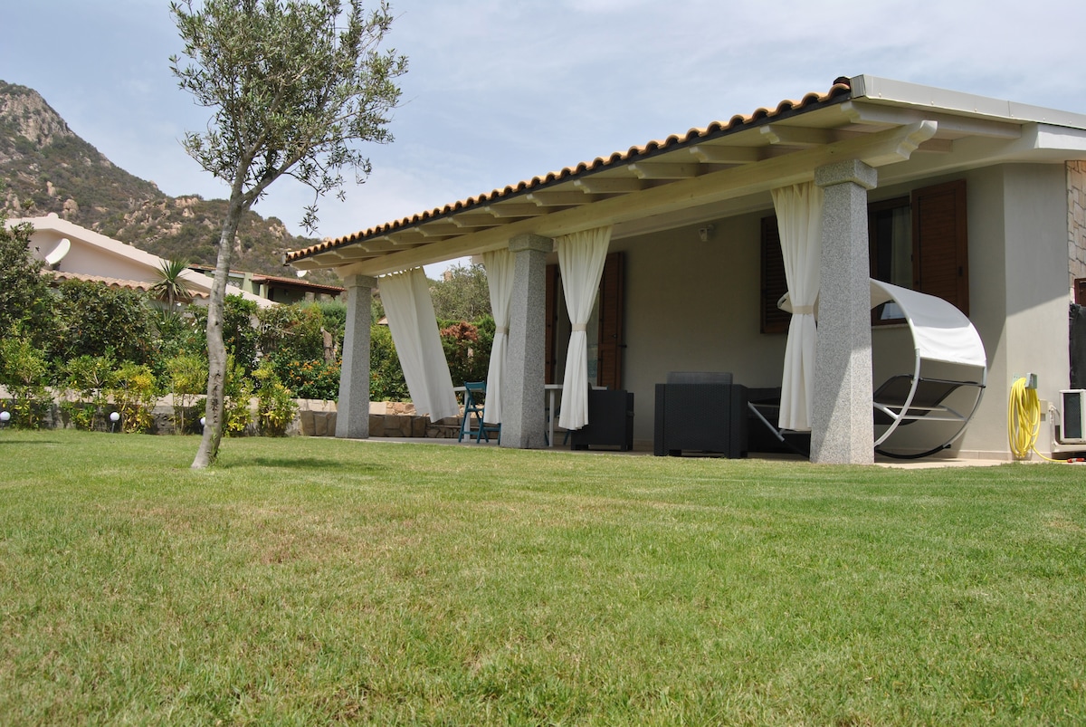 A single-story villa is set in a lush garden, featuring a covered veranda with elegantly draped curtains. A modern lounge area is visible, complemented by a hanging chair. Green grass surrounds the property, with cultivated plants and a gentle hill in the distance.