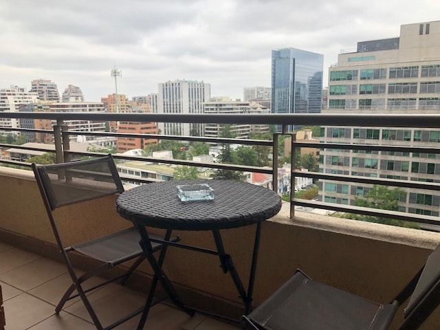 A balcony view showcases a mix of modern buildings and greenery in Santiago. Two folding chairs flank a small round table, accompanied by a glass ashtray. The railing offers a clear line of sight to the cityscape beyond, under a slightly overcast sky.