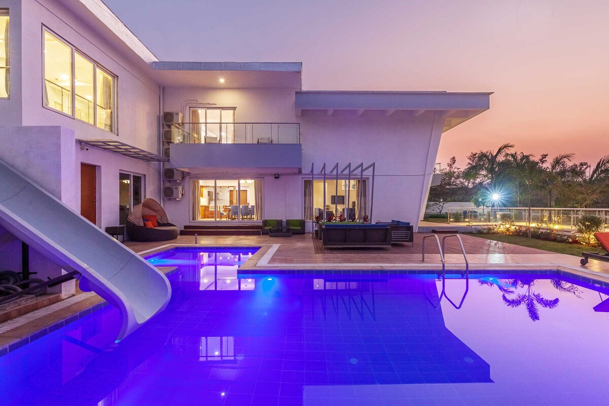 The villa's modern exterior is complemented by a spacious pool, featuring a slide that extends towards the shimmering water. The setting sun casts a soothing glow, highlighting the outdoor seating area and large windows of the living space visible in the background.