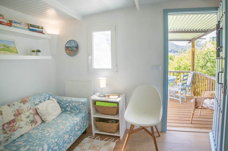 A bright lounge area features a floral-patterned couch and a small table with a lamp. Nearby, a window provides access to a wooden deck setting with outdoor seating, allowing views of the surrounding nature.
