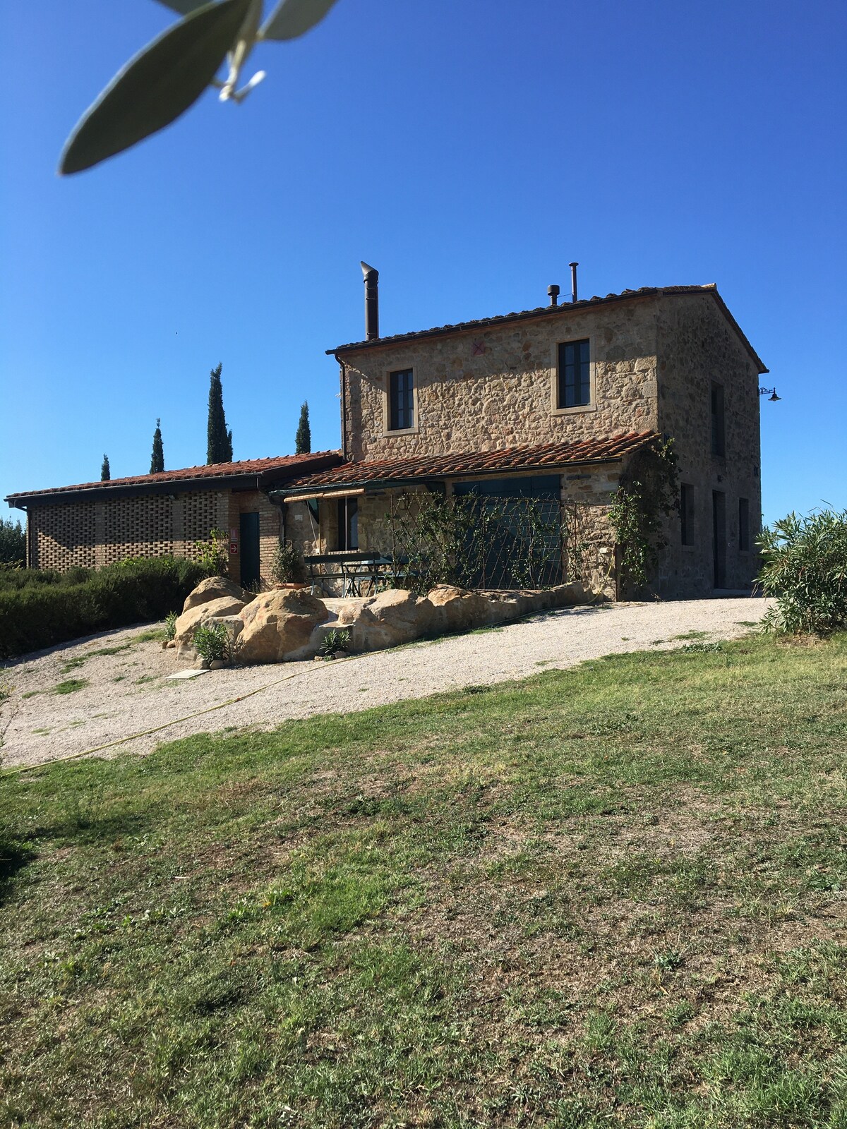 A two-story stone farmhouse is set against a clear blue sky, showcasing a rustic charm. The exterior features a mix of natural stone and terracotta elements, with a small terrace and garden area surrounded by greenery. A pathway leads up to the entrance.