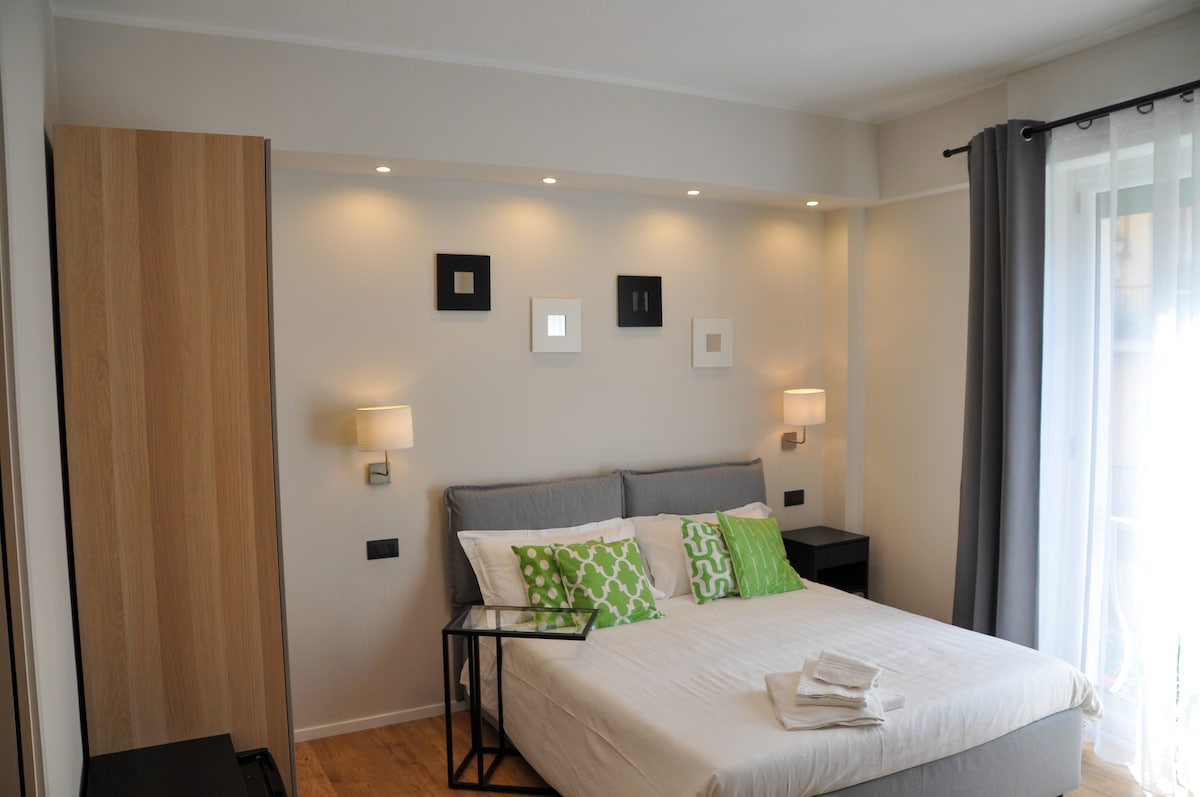 A comfortable bedroom features a spacious bed adorned with geometric patterned pillows. Soft lighting is provided by bedside lamps mounted on the walls, and artwork is displayed above the bed. Large windows allow natural light to fill the room, complemented by elegant curtains.