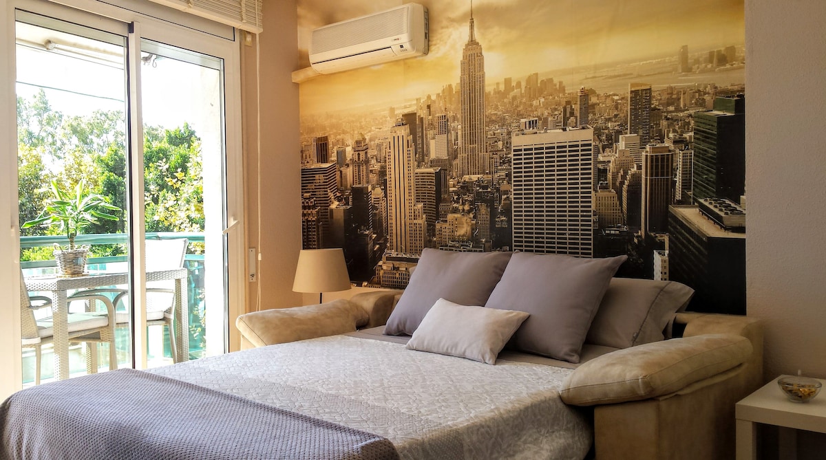 A spacious area features a comfortable sofa bed adorned with soft pillows. Light-colored linens cover the bed, complemented by a decorative throw. A large window allows natural light to fill the room, with a city skyline mural creating a striking backdrop. A small table and lamp add warmth.