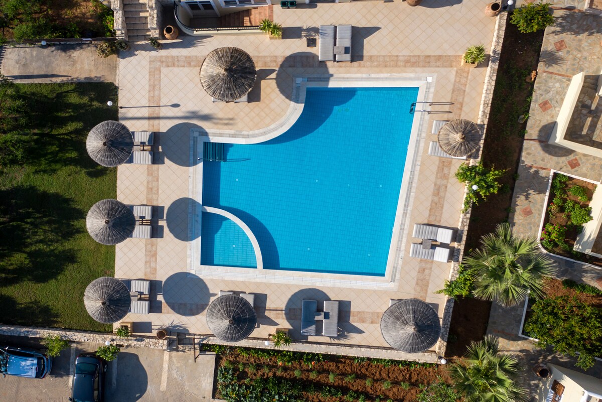 An aerial view displays a large swimming pool surrounded by equipped sun loungers and parasols. The pool's vibrant blue water contrasts with the neutral tiled deck. Lush greenery and tasteful landscaping can be seen surrounding the pool area, enhancing the welcoming atmosphere.