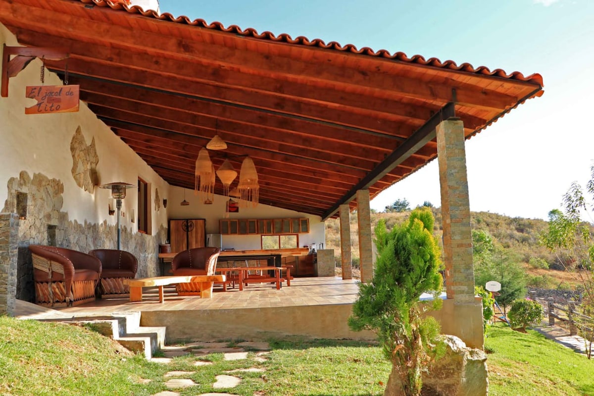 An expansive outdoor terrace features a wooden roof supported by stone columns. Comfortable seating arrangements are visible, accompanied by a large dining table. Lush greenery surrounds the area, with natural hills visible in the background under a clear blue sky.