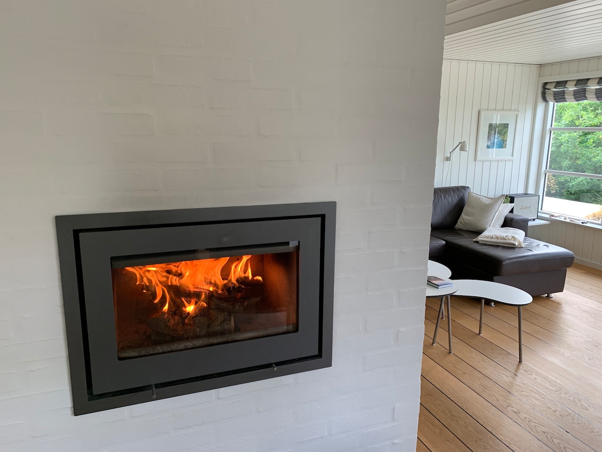 A built-in fireplace is positioned within a white brick wall, featuring a warm fire that adds a cozy element to the living space. In the background, a spacious seating area is visible with a dark sofa, complemented by a small coffee table and large windows that allow natural light.