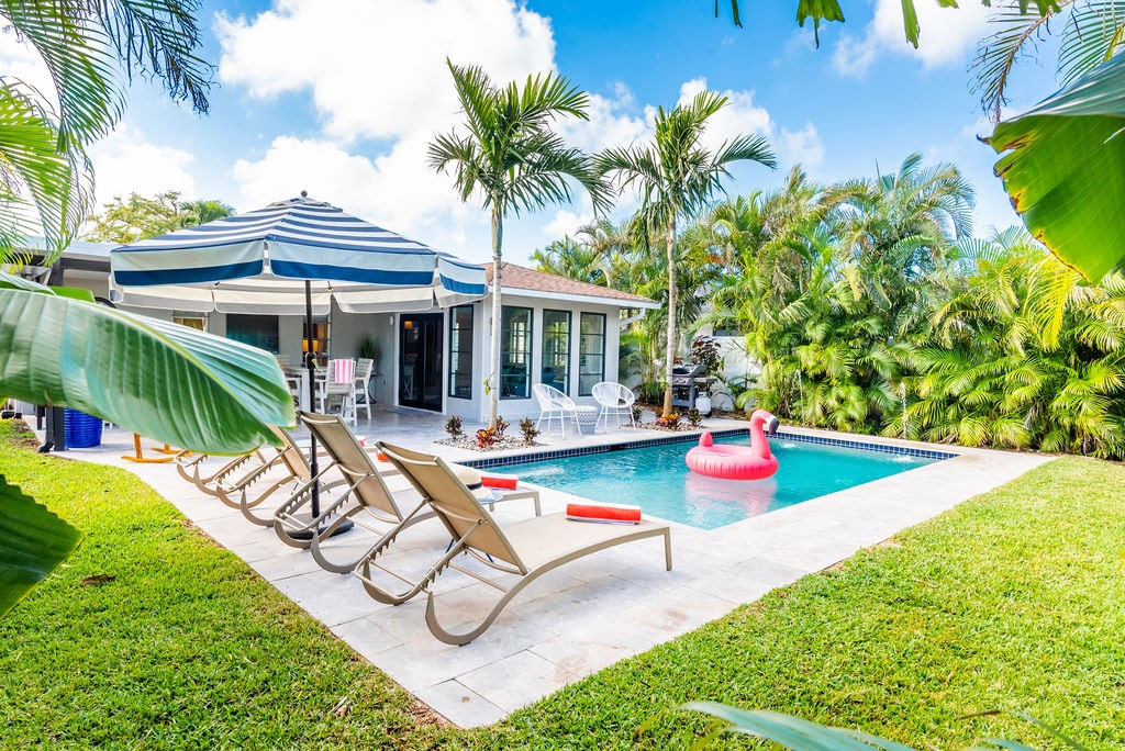 A backyard oasis is presented, featuring a heated pool bordered by tropical landscaping. Lounge chairs are arranged near the pool, complementing a large striped umbrella. A pink flamingo float adds a playful touch to the inviting water, with a spacious patio area visible in the background.