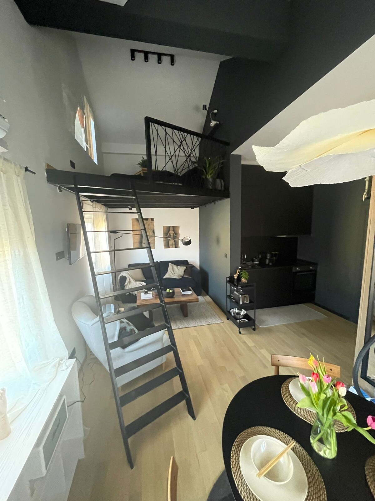 A sleek studio apartment is captured, featuring a cozy lounge area with a modern sofa and coffee table. A ladder leads to the elevated sleeping area, which includes a built-in projector. Natural light filters through sheer curtains, highlighting the minimalist decor and dark kitchen cabinetry.