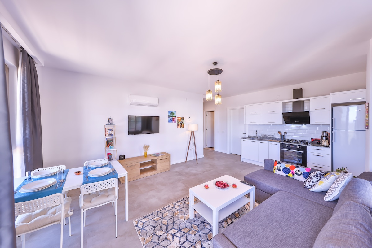 An open living area features a spacious gray sofa adorned with decorative pillows. A modern dining table is set with blue placemats, accompanied by light-colored chairs. A flat-screen TV is mounted on the wall, and a well-equipped kitchen is visible in the background.