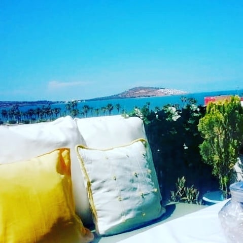 A cozy seating area features soft cushions in white and yellow, set against a backdrop of a clear blue sky and tranquil sea. Lush greenery surrounds the space, complementing the splendid ocean view and creating a serene atmosphere for relaxation.