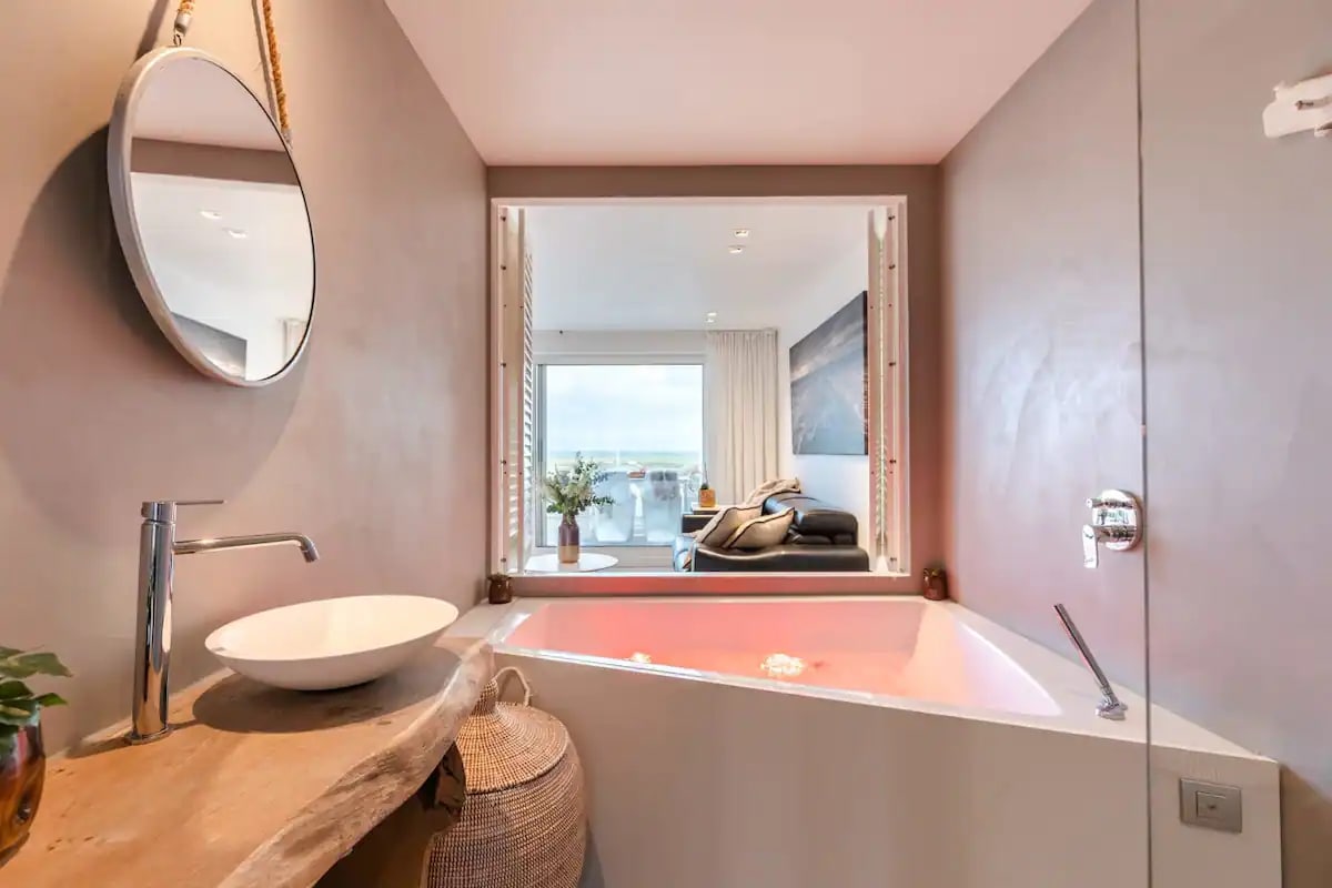 A modern bathroom features a sculptural bathtub with soft lighting. A round mirror reflects natural light, while a glimpse of the living area is visible through large windows. Soft neutral tones create a serene ambiance, enhancing the bathing experience.