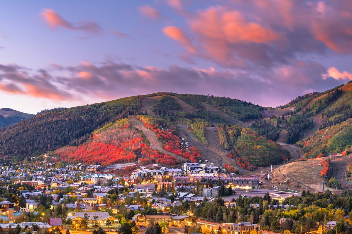 A panoramic view captures a colorful autumn scene in Park City, with vibrant hues of red and orange covering the mountainside. The serene landscape extends over the town and ski area, with soft clouds drifting across a pastel sky at dusk.