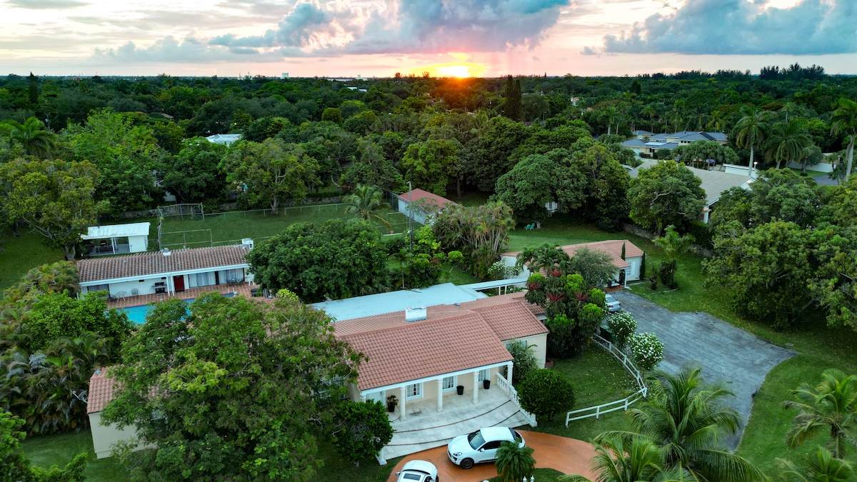 An aerial view showcases the sprawling property, with lush greenery surrounding the cottage and pool area. A vibrant sunset casts warm hues over the landscape, while the layout of the ranch and parking space is clearly visible. Tall trees accentuate the tranquil outdoor setting.