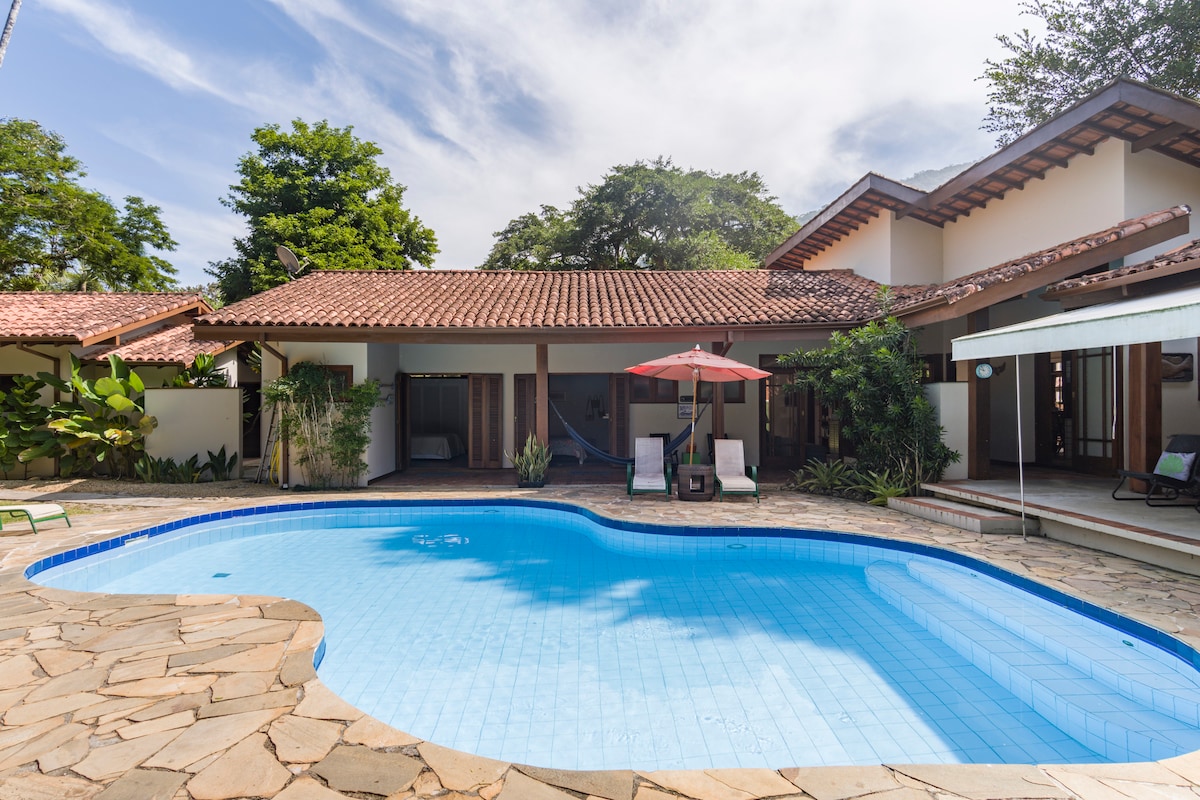 A clear swimming pool is set amid a well-maintained outdoor space, featuring a natural stone patio. Lounge chairs and an umbrella are positioned nearby, surrounded by lush greenery and shaded by trees. The house's roof tiles complement the tranquil ambiance of the area.