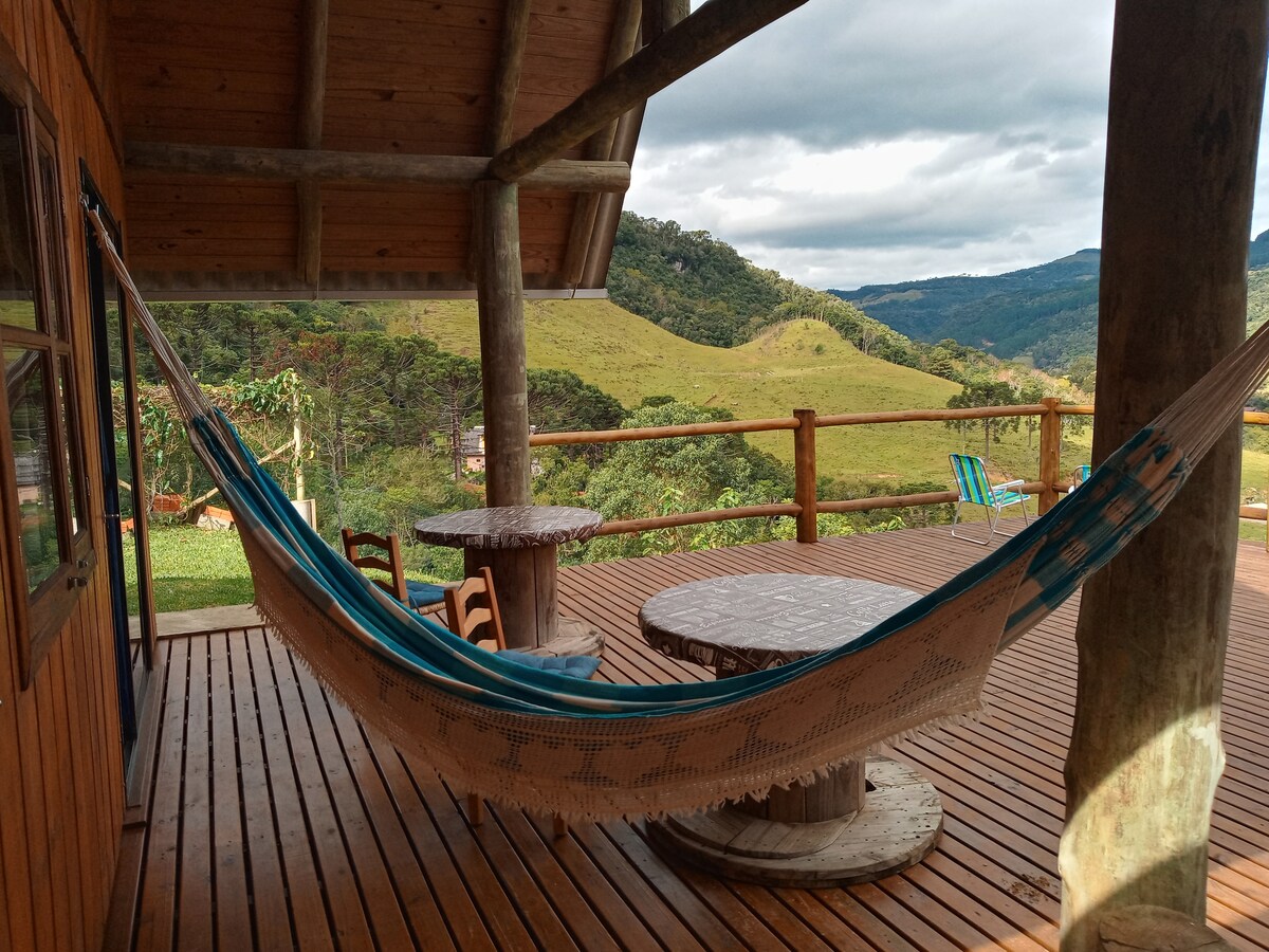 A spacious deck features a hammock and a round wooden table surrounded by chairs. Expansive views of green hills and trees are visible in the background under a partly cloudy sky.