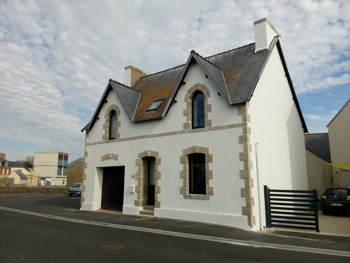 A two-story house features a white exterior with stone accents and multiple arched windows. The roof is covered with dark tiles, and a skylight is visible. The entrance has a large archway, and a gated pathway leads to the house, situated along a quiet street.