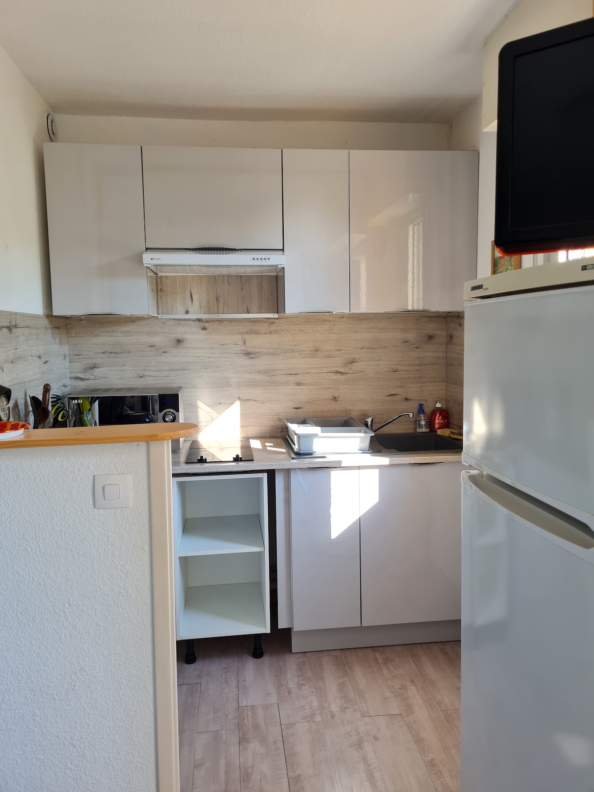 A modern kitchen space features sleek cabinetry and a combination of appliances, including a refrigerator and a microwave. Natural light filters through a window, illuminating the work area, which is equipped with essential utensils and a small stove beneath the exhaust hood.