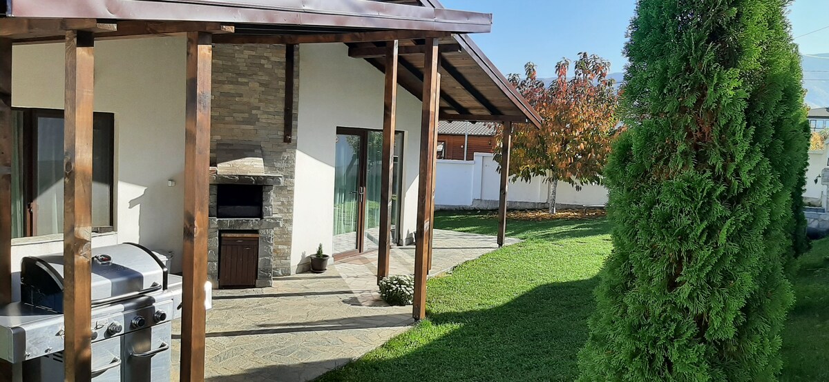 An outdoor space features a patio with a barbecue grill and stone chimney. The area is surrounded by well-maintained grass and has two trees, providing shade. The modern house is visible in the background, showcasing large windows and a sleek roofline.