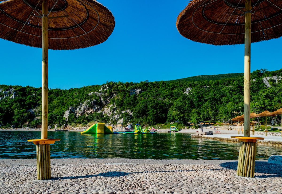 Two thatched umbrellas are positioned over wooden stands on a pebbled beach. In the background, lush greenery lines the shore, while bright slides are visible in the water. The clear blue sky complements the tranquil scene, inviting relaxation by the gentle waves.
