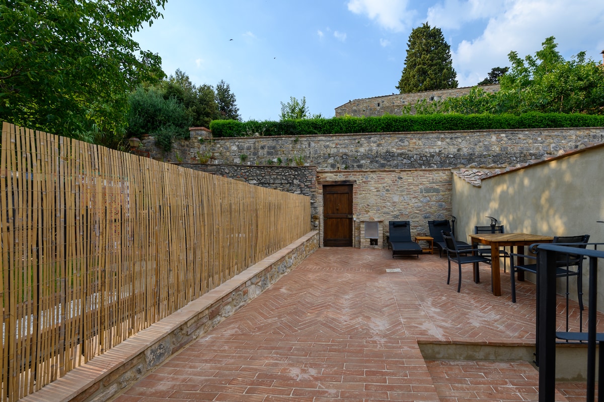 An outdoor courtyard features a stone wall lined with greenery, providing a serene setting. Comfortable lounge chairs and a wooden table are placed on the terracotta tiles, creating a space for relaxation or dining. The area is enclosed for privacy and tranquility.
