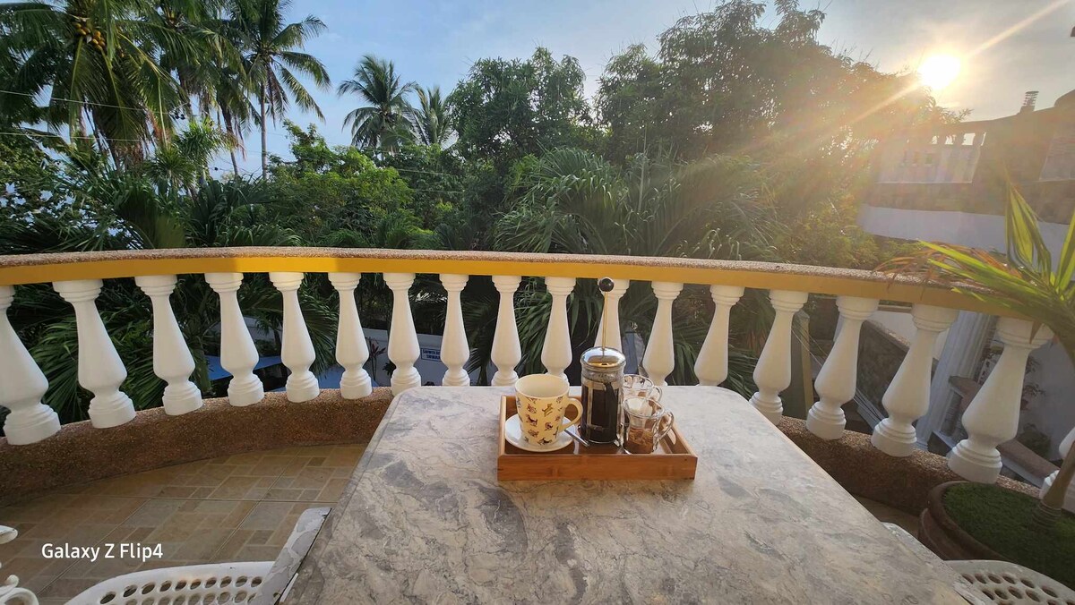 A balcony is showcased with a marble-topped table holding a wooden tray containing coffee and pastries. Lush tropical foliage surrounds the outdoor space, while the sun rises in the background, casting a warm light over the scene.