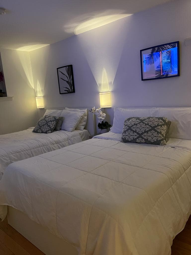 A bright bedroom features two spacious beds with white bedding and decorative pillows. Soft lighting is provided by wall-mounted lamps, creating a serene environment. Framed artwork is displayed on the walls, complemented by light-colored flooring that enhances the room's openness.