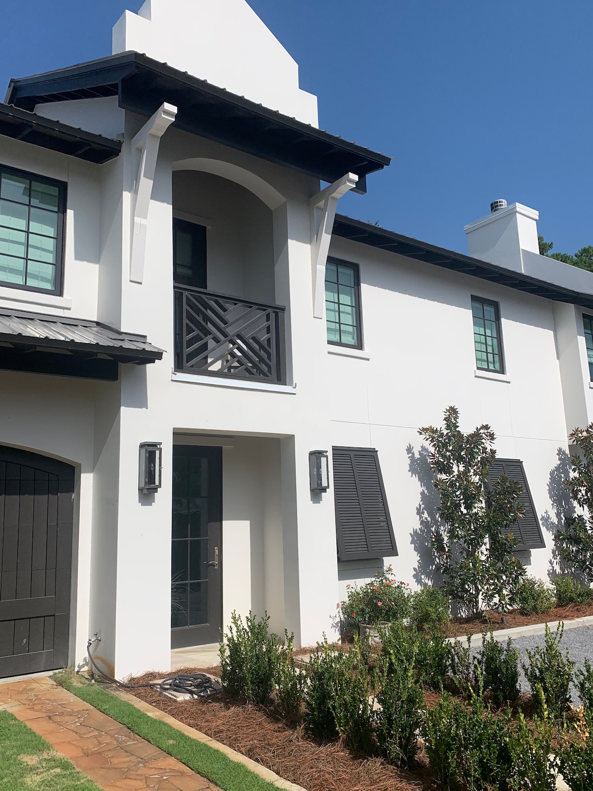 A modern exterior view of the carriage house showcases crisp white walls and dark accents. A small balcony with a decorative railing is visible above the entrance, framed by two windows. Neatly trimmed shrubs line the pathway, leading to a black door that complements the architecture.