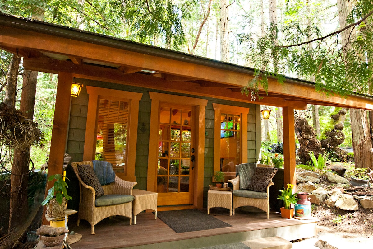 The cabin's exterior features a welcoming porch with two wicker chairs. Surrounded by tall trees, warm light fixtures enhance the inviting entrance, which includes double wooden doors. Lush greenery and thoughtfully arranged natural elements create a serene atmosphere.