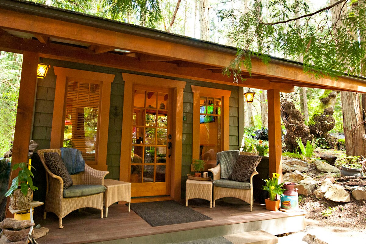 The cabin entrance is framed by a spacious porch featuring two comfortable chairs. A welcoming wooden door with glass panels leads inside, flanked by warm lighting and surrounded by lush greenery. Natural textures and colors complement the serene forest setting.