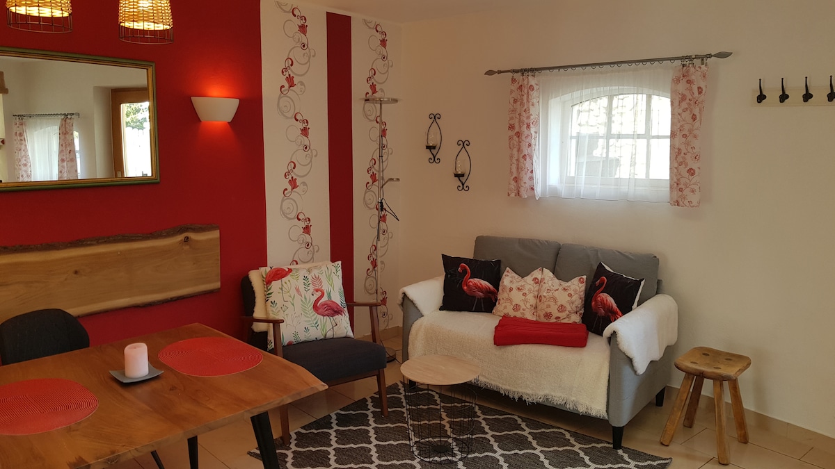 A living area is showcased with a neutral sofa adorned with decorative pillows featuring flamingos. A wooden dining table is set nearby, with round placemats placed on top. A mirror and wall decorations are visible, adding character, while natural light filters through the window with floral curtains.
