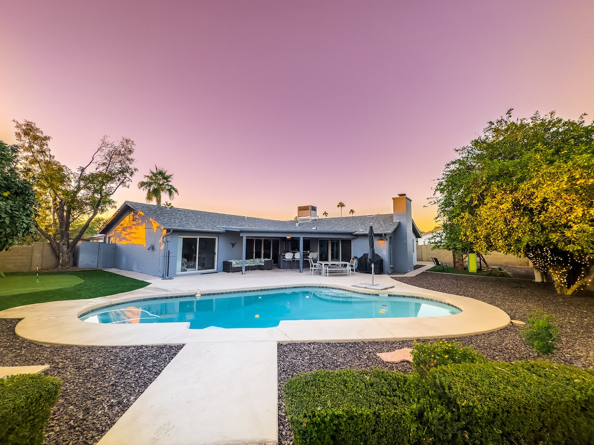 The exterior of the home is presented with a large pool surrounded by landscaped areas featuring shrubs. The structure's modern design is highlighted against a twilight sky, and windows reflect the warm glow from within.
